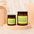 Two amber glass candles with black lids and "Here's to Forever" labels.