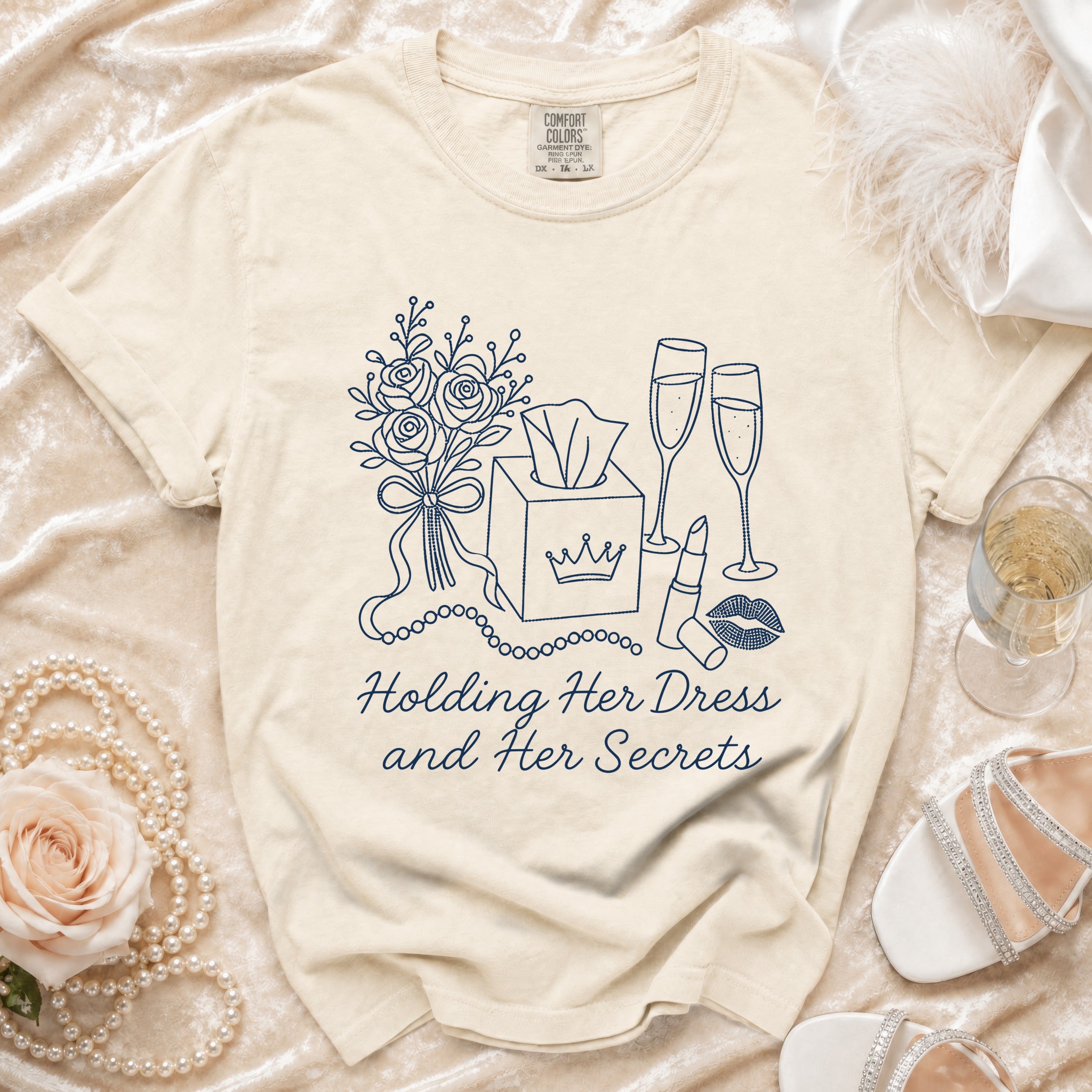 Cream t-shirt with navy line art of roses, tissue box, champagne, and lipstick. Text reads "Holding Her Dress and Her Secrets."