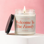 Glass jar candle with lit wick, black lid off to the side, reads "Welcome To The Family".