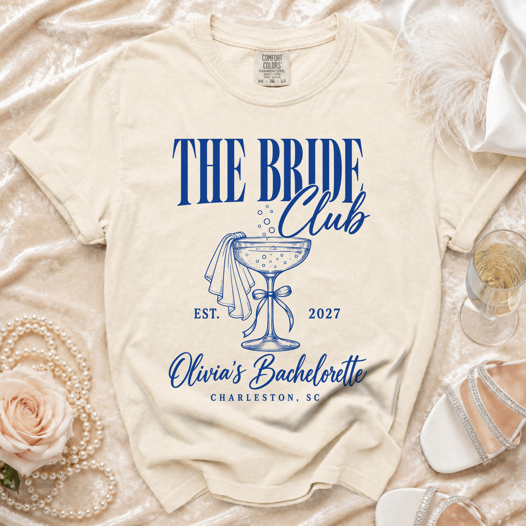 Cream t-shirt with blue text "The Bride Club", a champagne glass, and "Olivia's Bachelorette Charleston, SC".