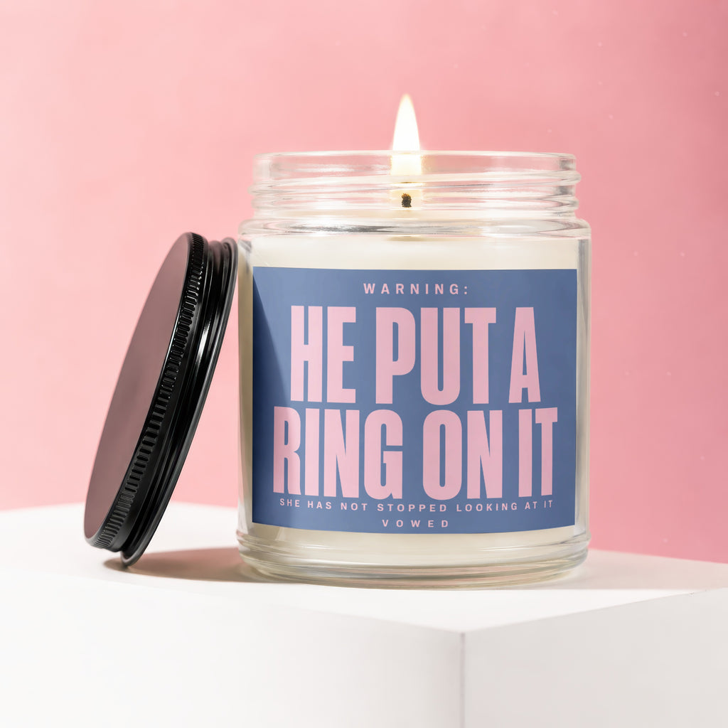 Candle with pink text "HE PUT A RING ON IT" on a blue label.
