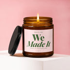 Amber glass candle with burning wick, lid off. Label reads "Smells Like We Made It, Against All Reasonable Odds".