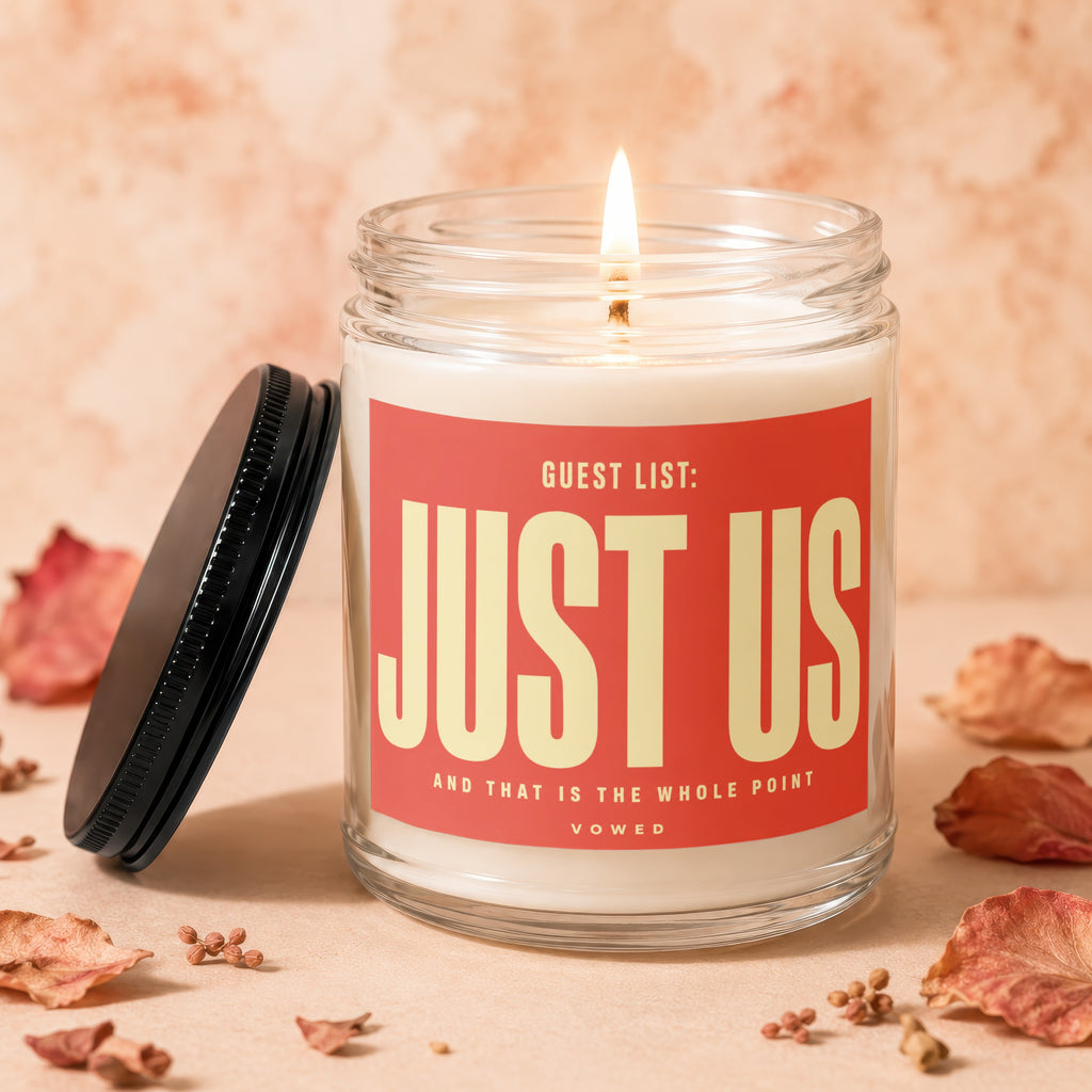 Glass jar candle with lit wick, red label reads "Guest List: JUST US"