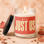 Glass jar candle with lit wick, red label reads "Guest List: JUST US"