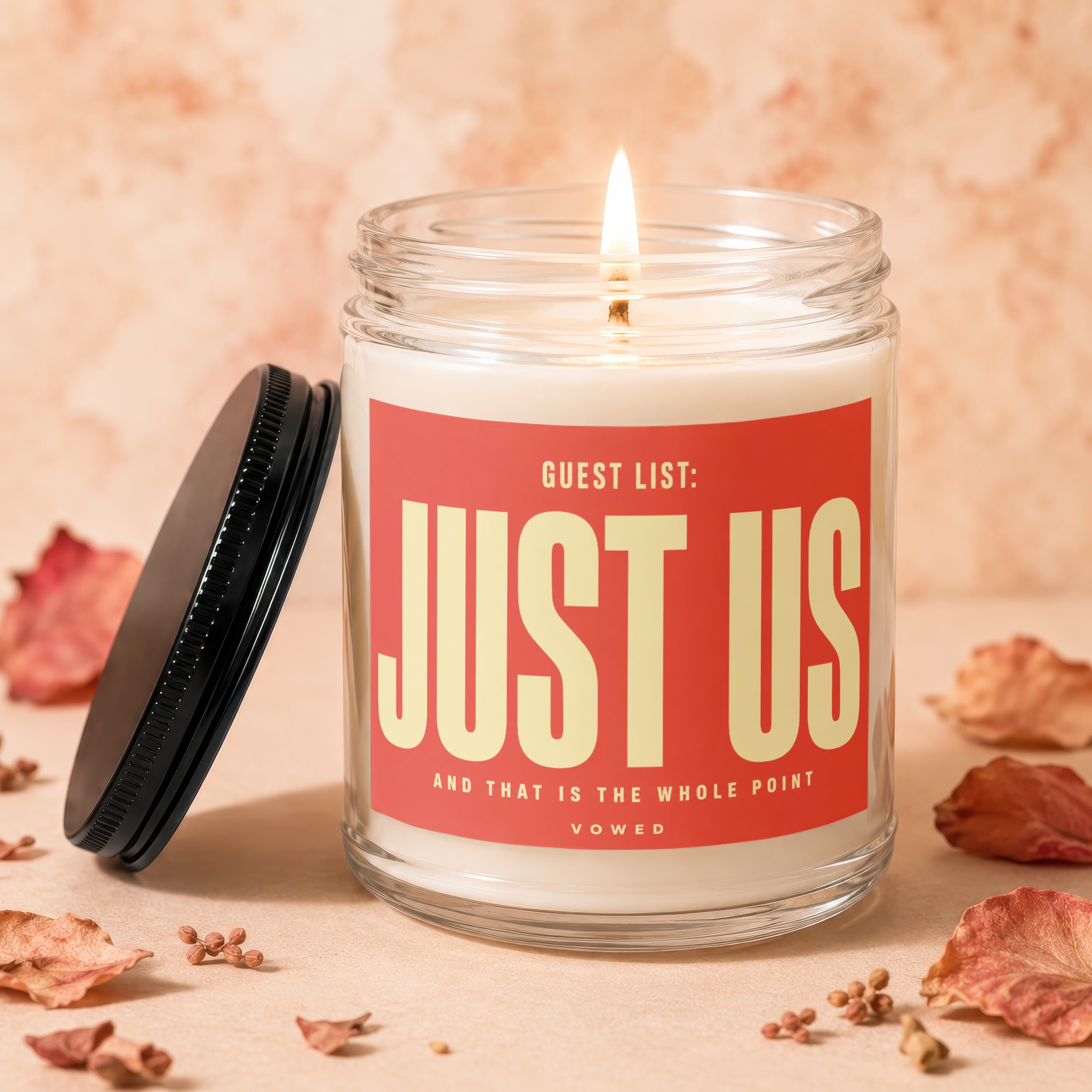 Glass jar candle with lit wick, red label reads "Guest List: JUST US"