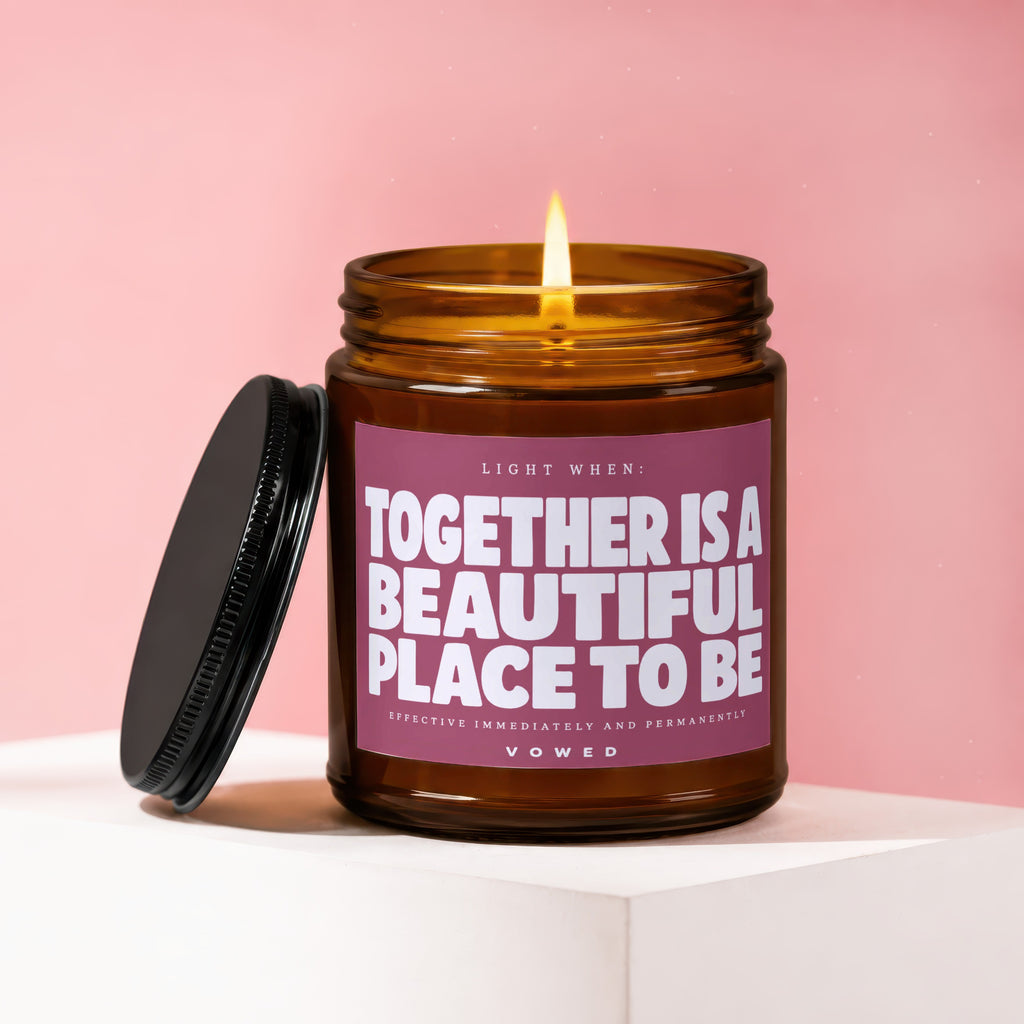 Amber jar candle with lit wick, black lid, and pink label reading "Together is a beautiful place to be."