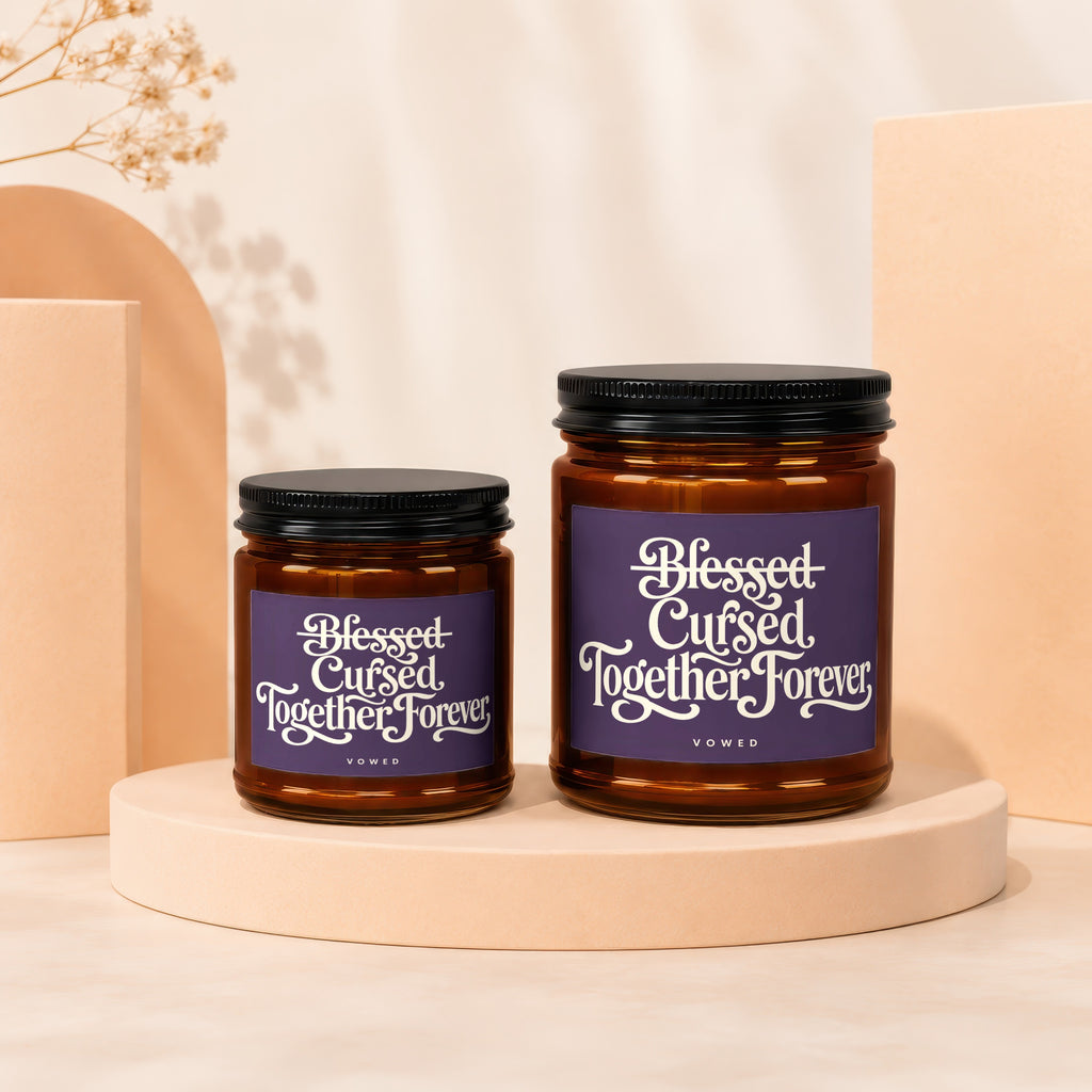 Two amber glass candles with black lids, labeled "Blessed Cursed Together Forever."