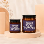 Two amber glass candles with black lids, labeled "Blessed Cursed Together Forever."