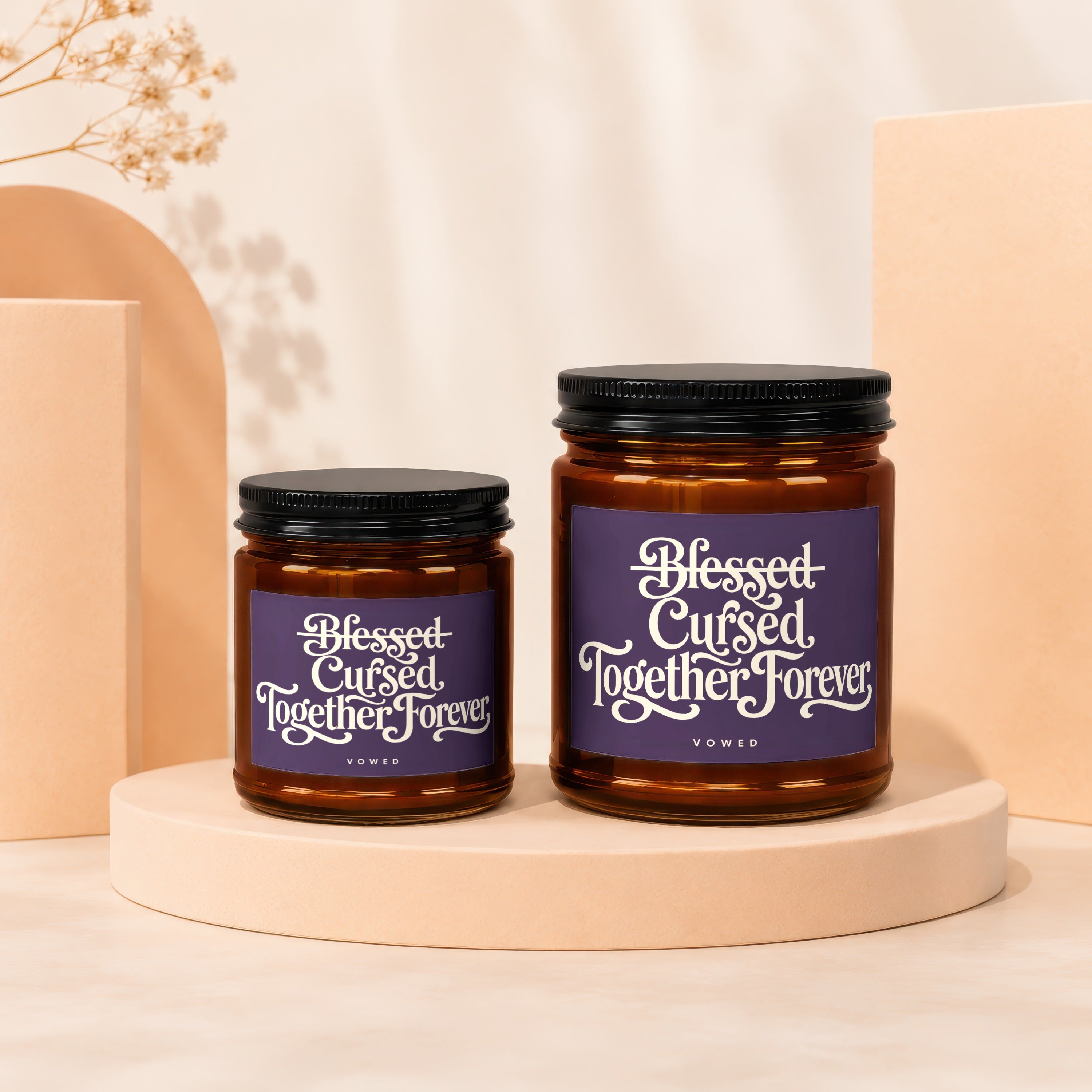 Two amber glass candles with black lids, labeled "Blessed Cursed Together Forever."