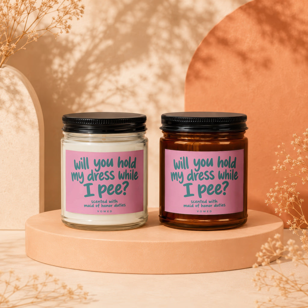 Two candles with pink labels reading "will you hold my dress while I pee?".
