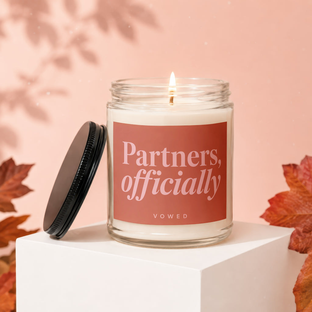 Candle labeled "Partners, officially" in a glass jar with lid off, surrounded by autumn leaves.
