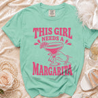 Teal t-shirt with pink text reading "THIS GIRL NEEDS A MARGARITA" and a graphic of a hand holding a margarita.