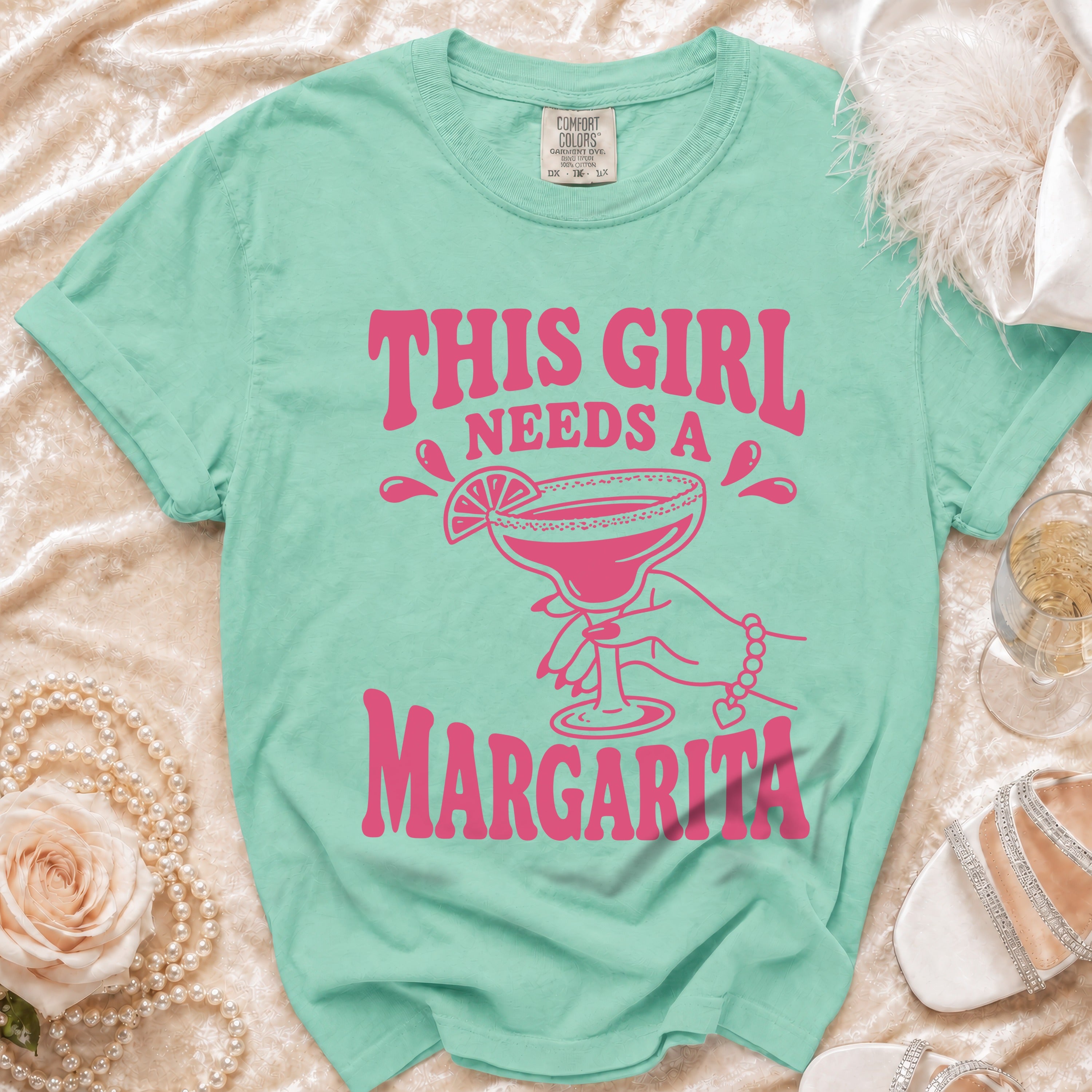 Teal t-shirt with pink text reading "THIS GIRL NEEDS A MARGARITA" and a graphic of a hand holding a margarita.