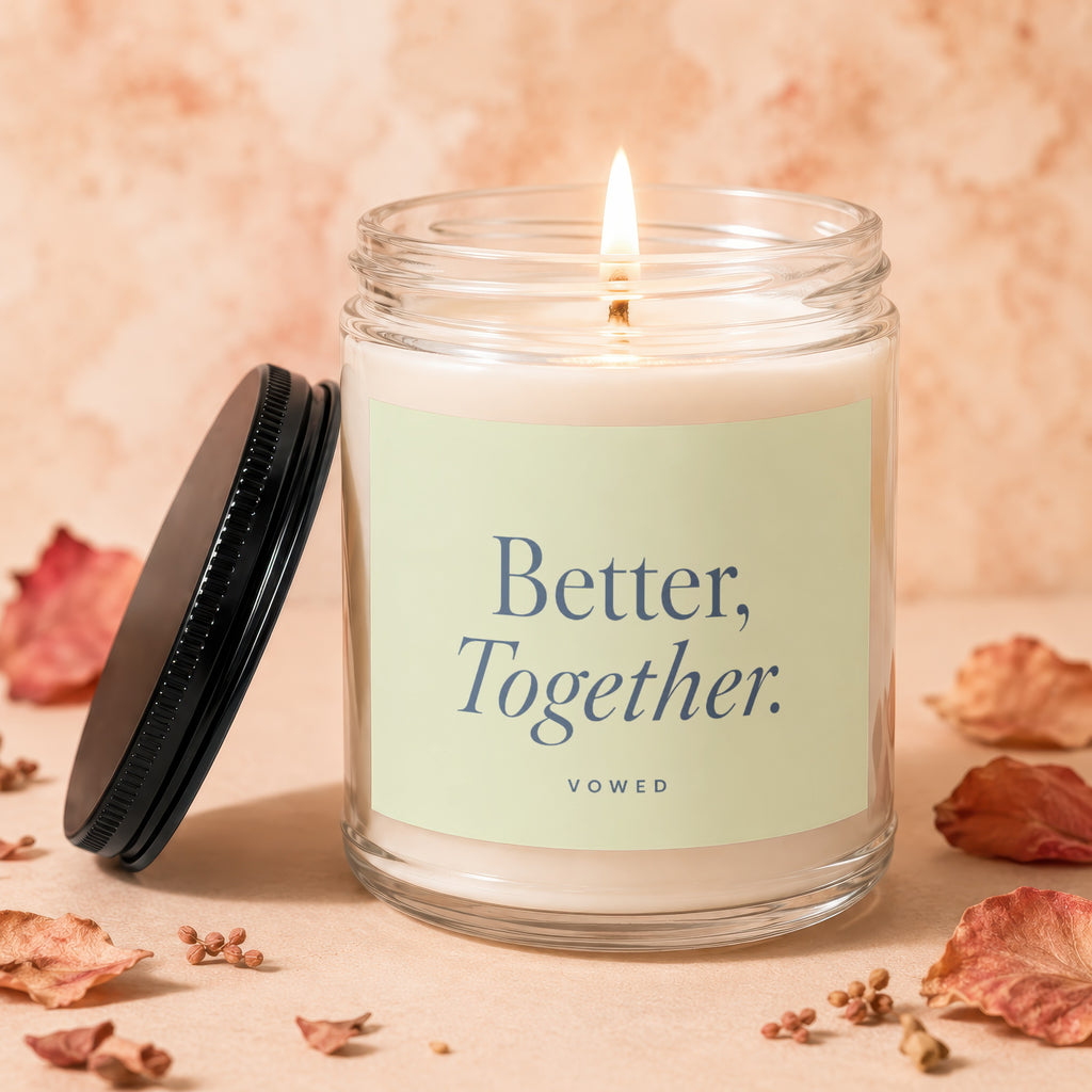 Lit glass candle with label "Better, Together. VOWED" surrounded by dried leaves.