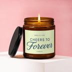 Amber jar candle with lit flame, "Cheers to Forever" text on label.