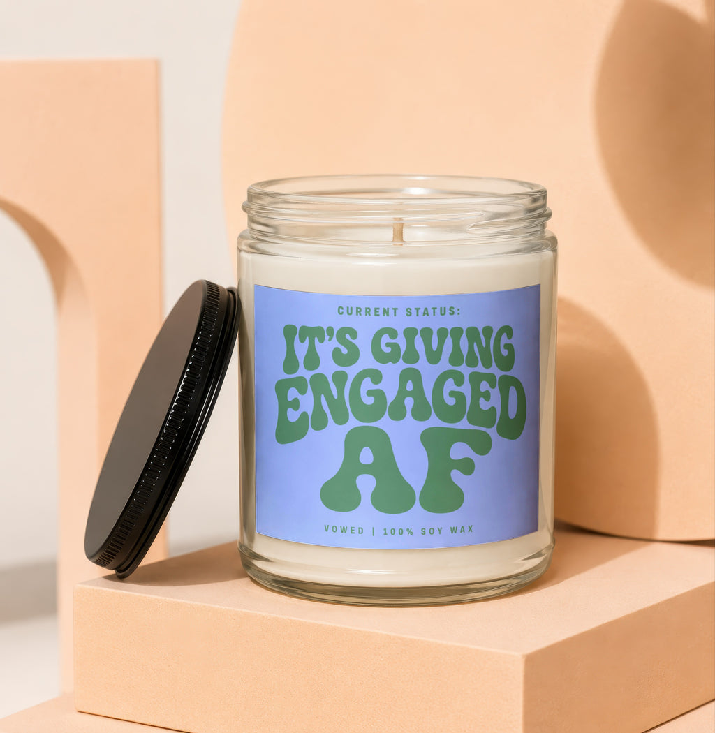 Glass candle with blue label reading "It's Giving Engaged AF" and "Vowed | 100% Soy Wax".