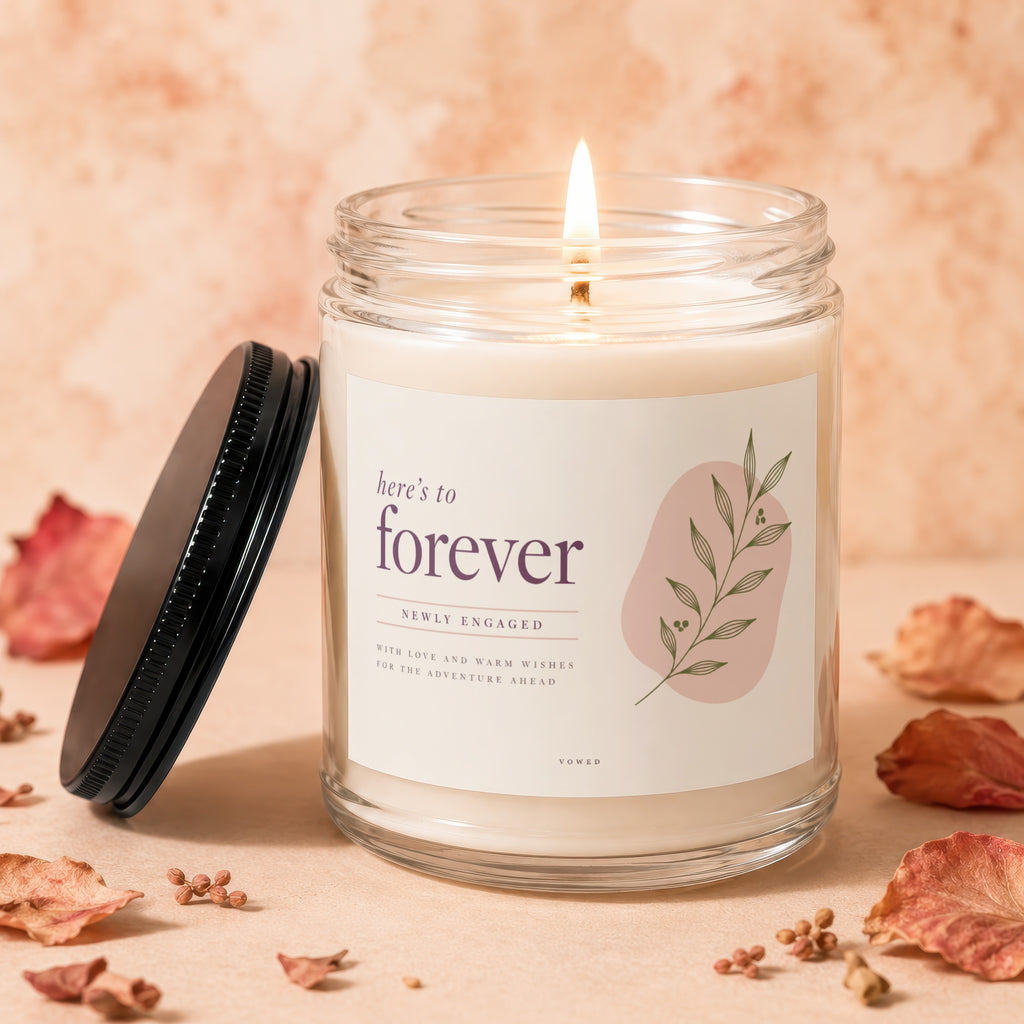 Engraved glass candle with lid off, flame lit, reading "here's to forever."