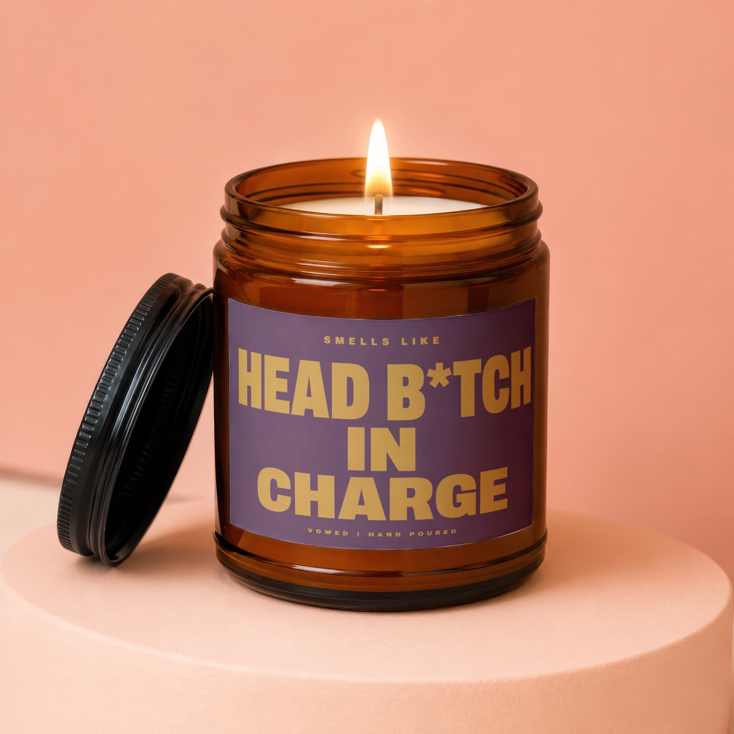 Amber jar candle with lit flame and purple label reading "SMFFLS LIKE HEAD B*TCH IN CHARGE".