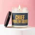 Glass candle with lit wick and dark label reading "TITLE: CHIEF PROBLEM SOLVER"