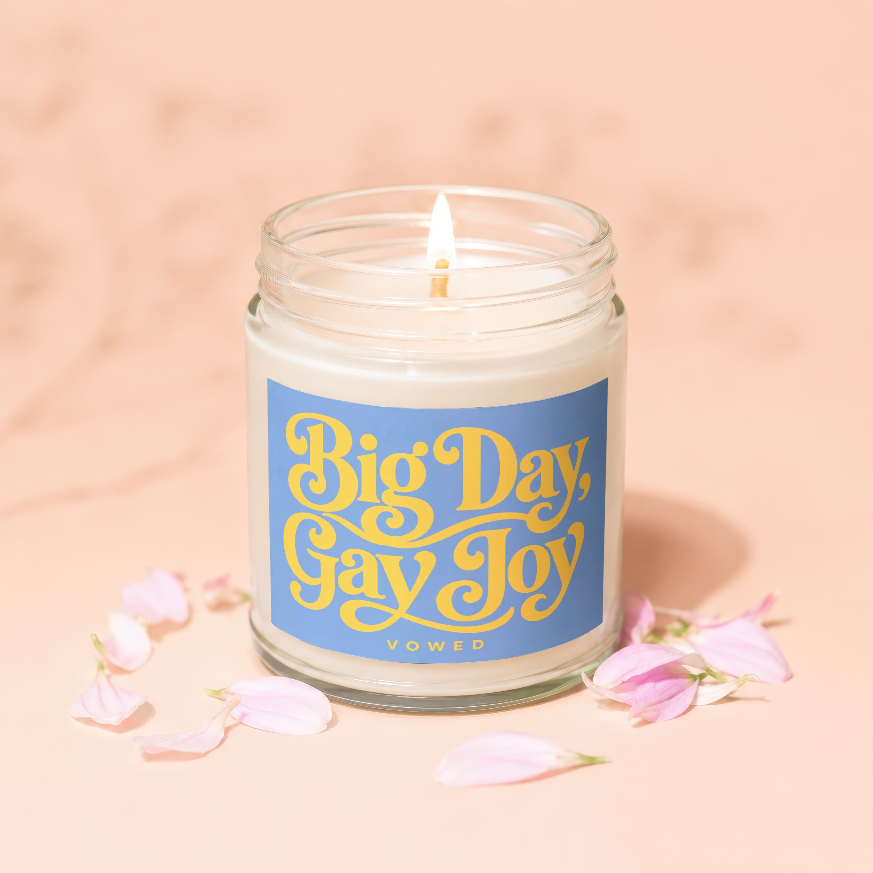 Glass jar candle with lit wick and blue label saying "Big Day, Gay Joy".