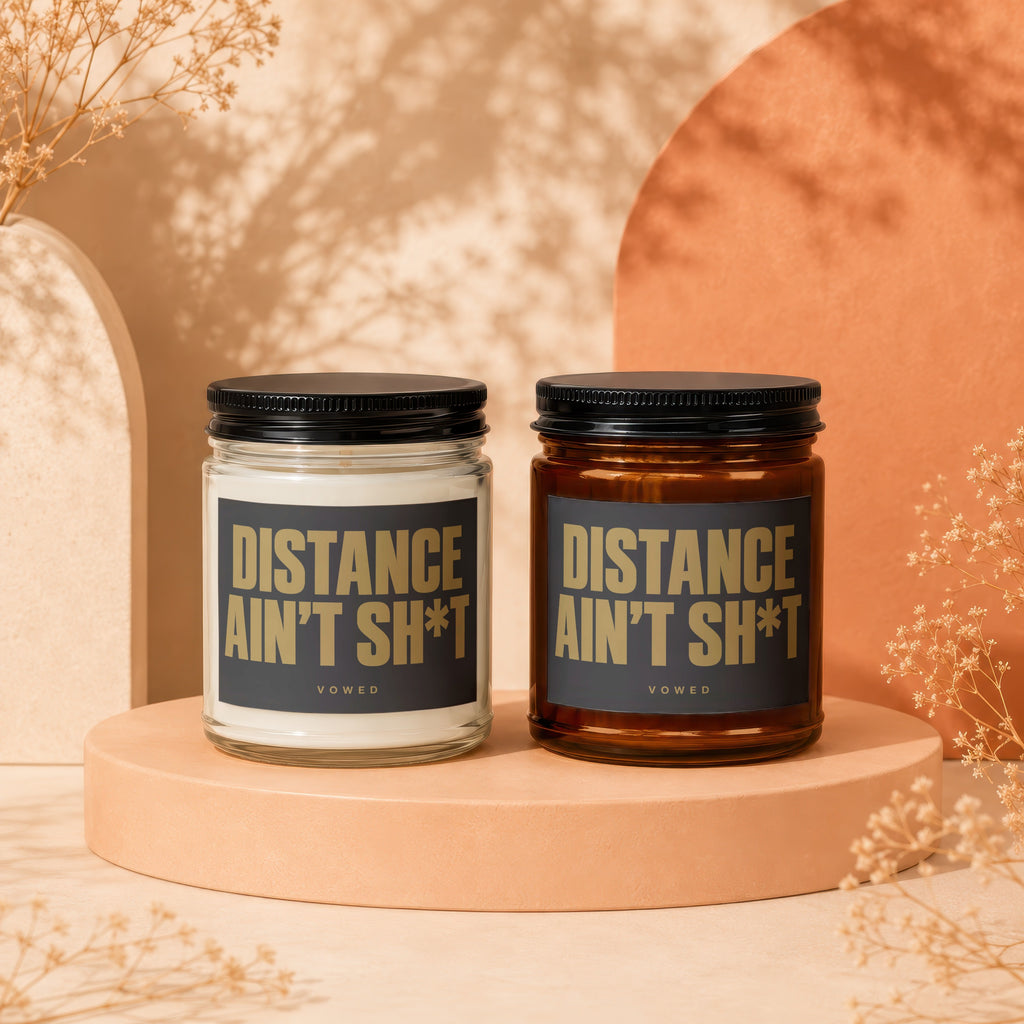Two candles with black lids, one clear and one amber jar, labeled "DISTANCE AIN'T SH*T".