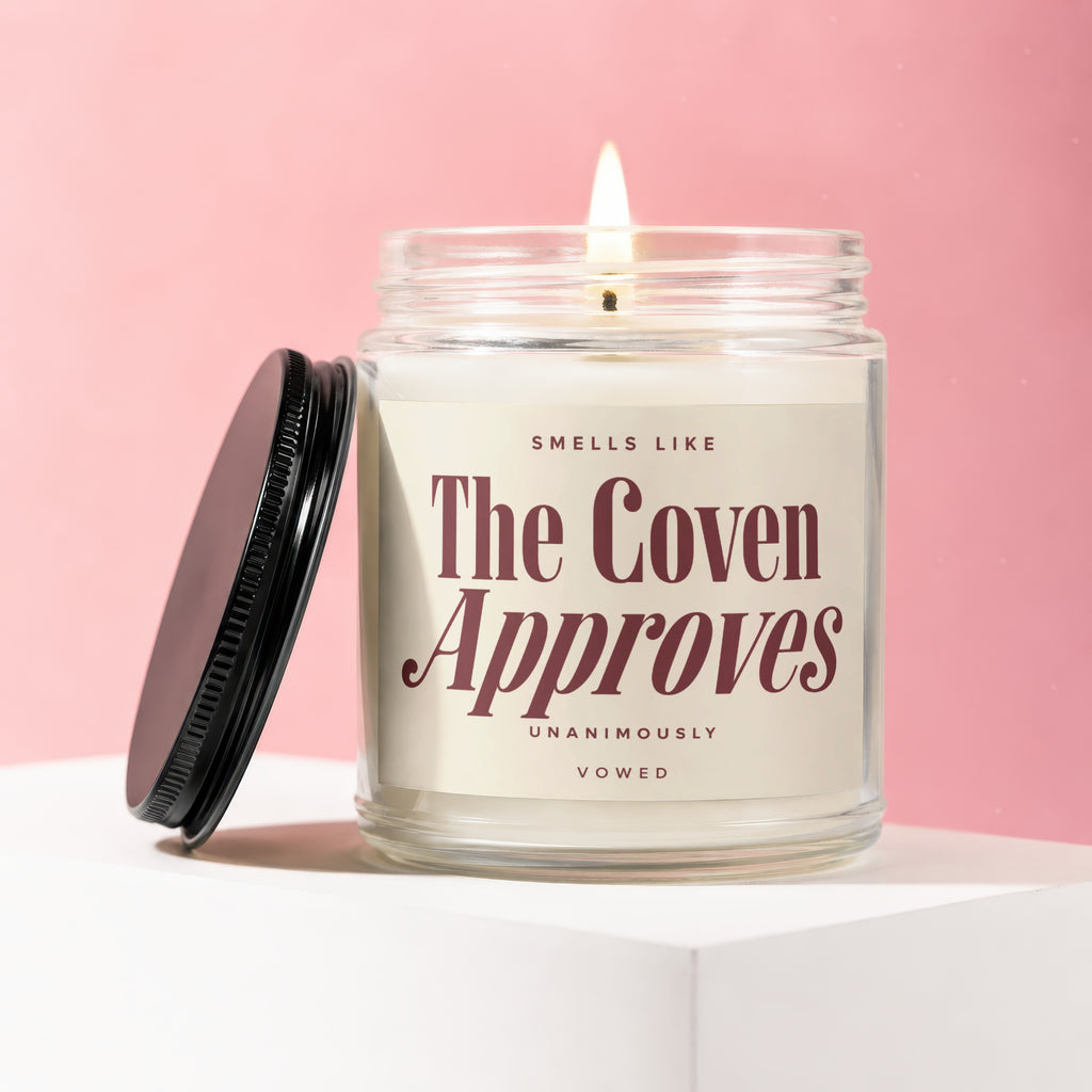 Glass jar candle with lit wick, reads "Smells like The Coven Approves".