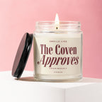 Glass jar candle with lit wick, reads "Smells like The Coven Approves".