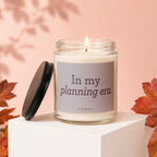 Glass candle with a lit wick on a white cube, reads "In my planning era."