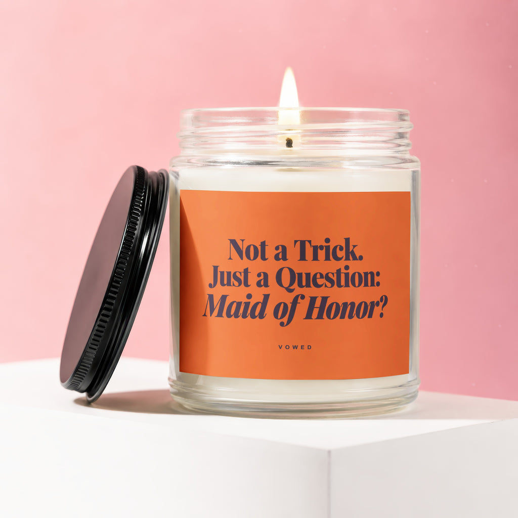 Glass candle with lit wick and orange label reading "Not a Trick. Just a Question: Maid of Honor?"