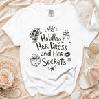 White t-shirt with "Holding Her Dress and Her Secrets" graphic, plus decorative elements.