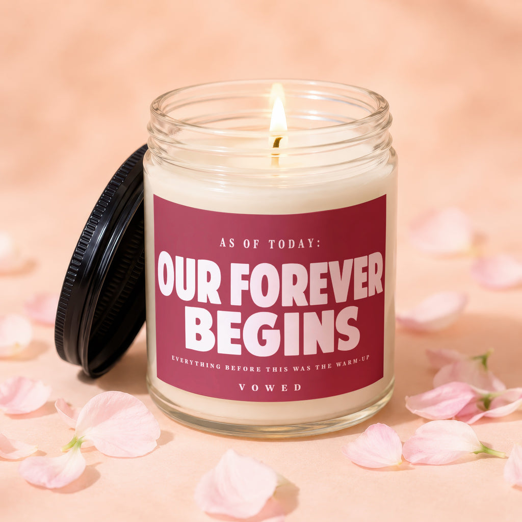 Candle with red label that reads "Our Forever Begins" surrounded by rose petals.