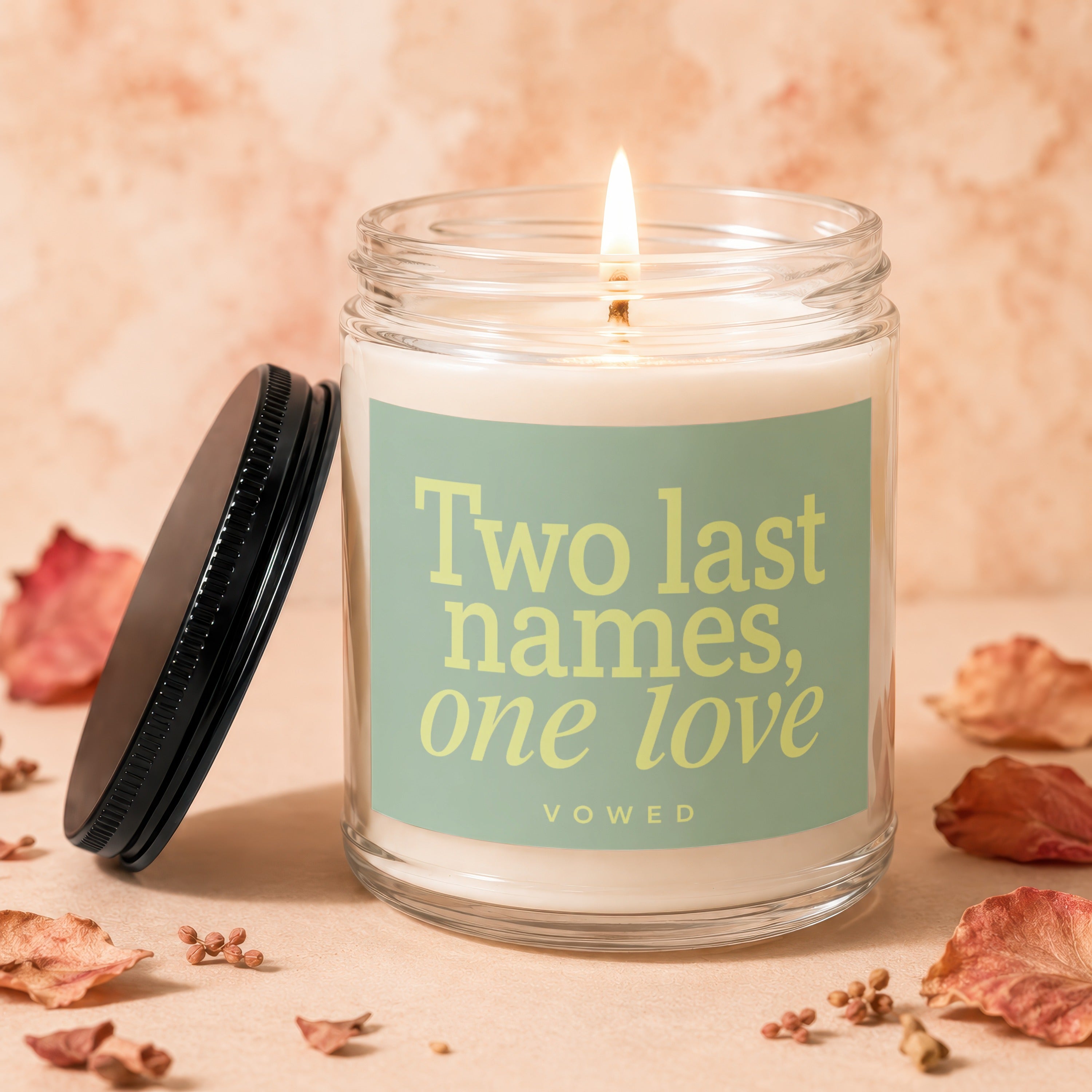 Glass candle with lit wick and label reading "Two last names, one love vōwed".