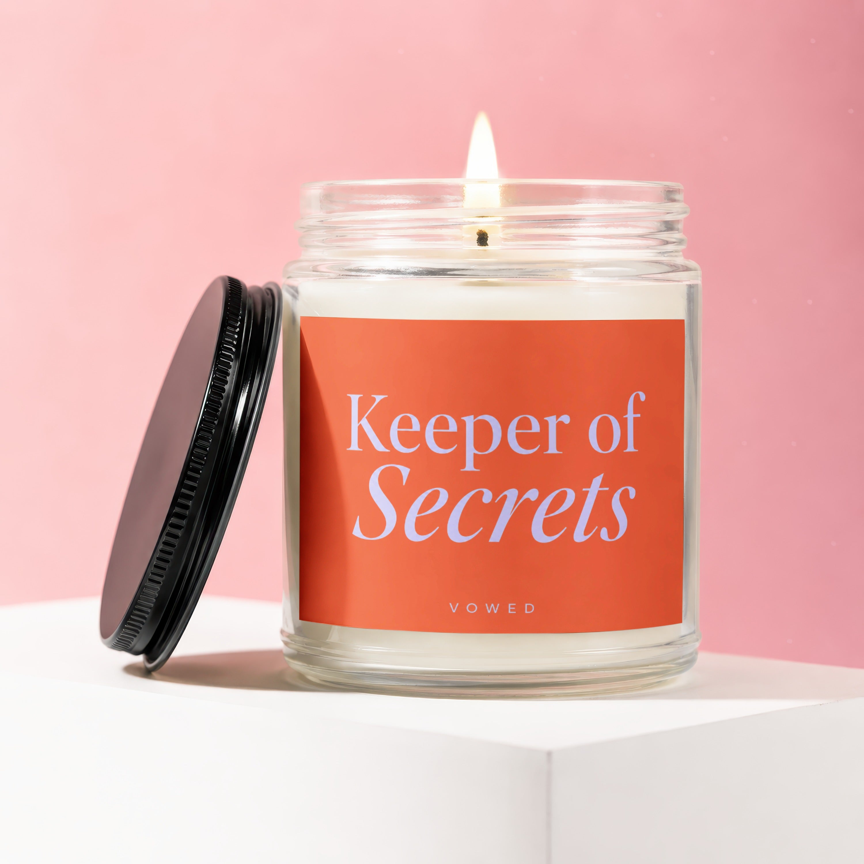 Glass jar candle with lit wick and orange label reading "Keeper of Secrets".
