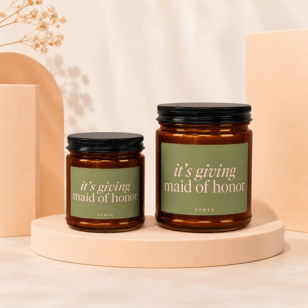 Two amber glass candles with green labels reading "it's giving maid of honor."