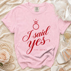 A pink Comfort Colors t-shirt with red text reading "I said yes THE BRIDE" and a diamond ring graphic, laid on a textured cream surface with accessories.