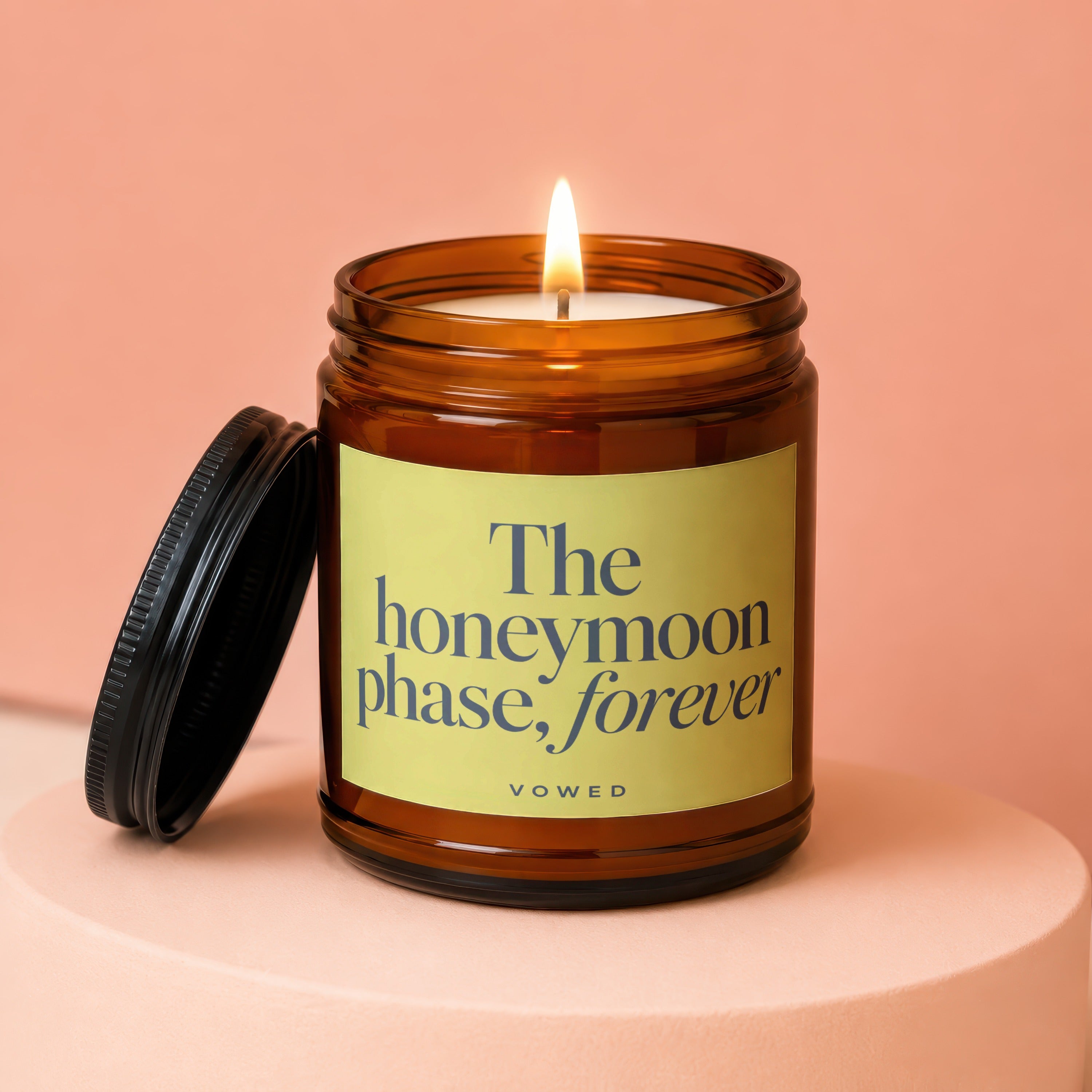 Amber jar candle with lit flame and black lid, reading "The honeymoon phase, forever"