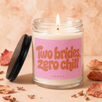 Glass jar candle with burning wick, pink label reading "Two brides, zero chill".