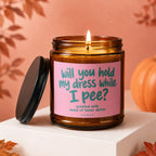 Glass candle with pink label asking "will you hold my dress while I pee?".