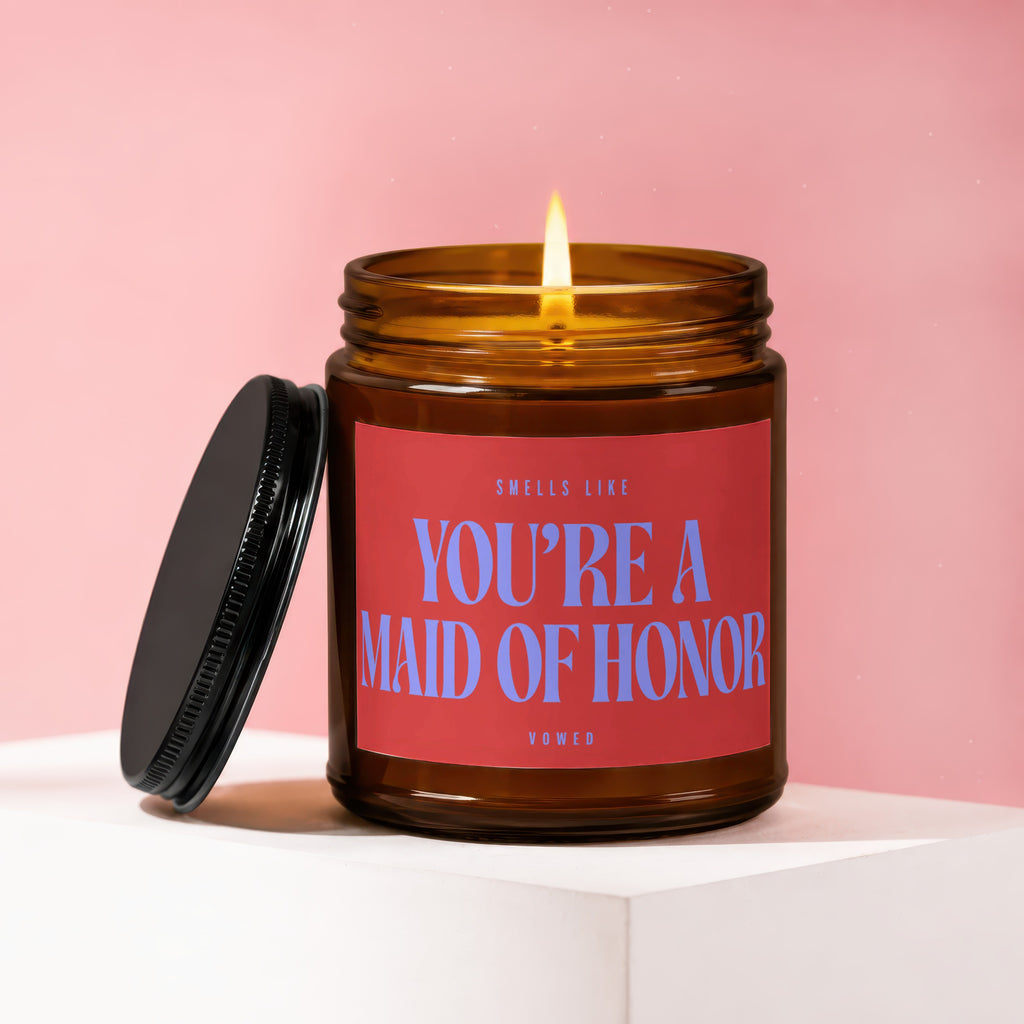 Amber glass candle with lit wick and red label reading "S pulls like You're a Maid of Honor Vowed"