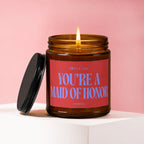 Amber glass candle with lit wick and red label reading "S pulls like You're a Maid of Honor Vowed"