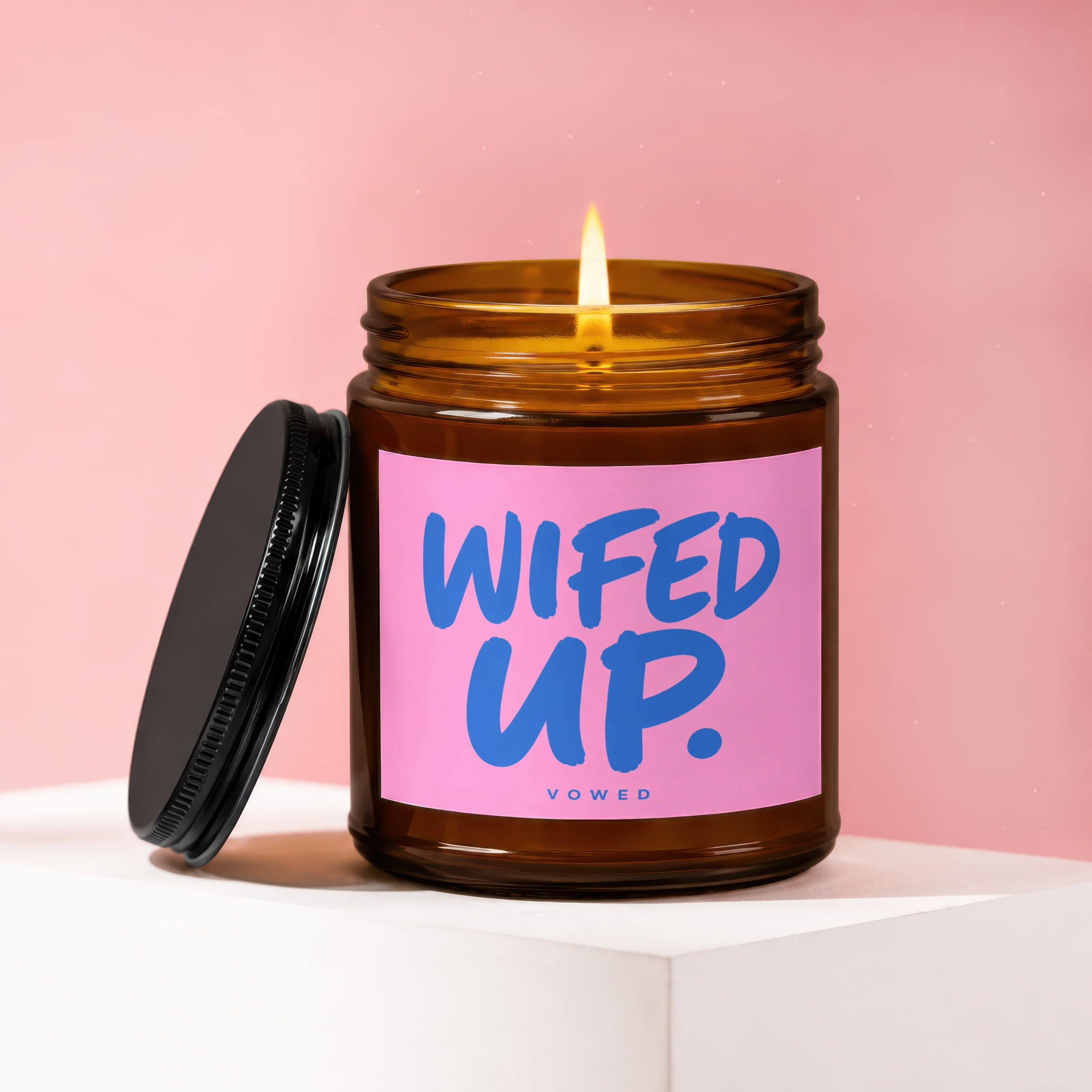 Amber glass candle with burning wick and pink label saying "WIFED UP. VOWED".