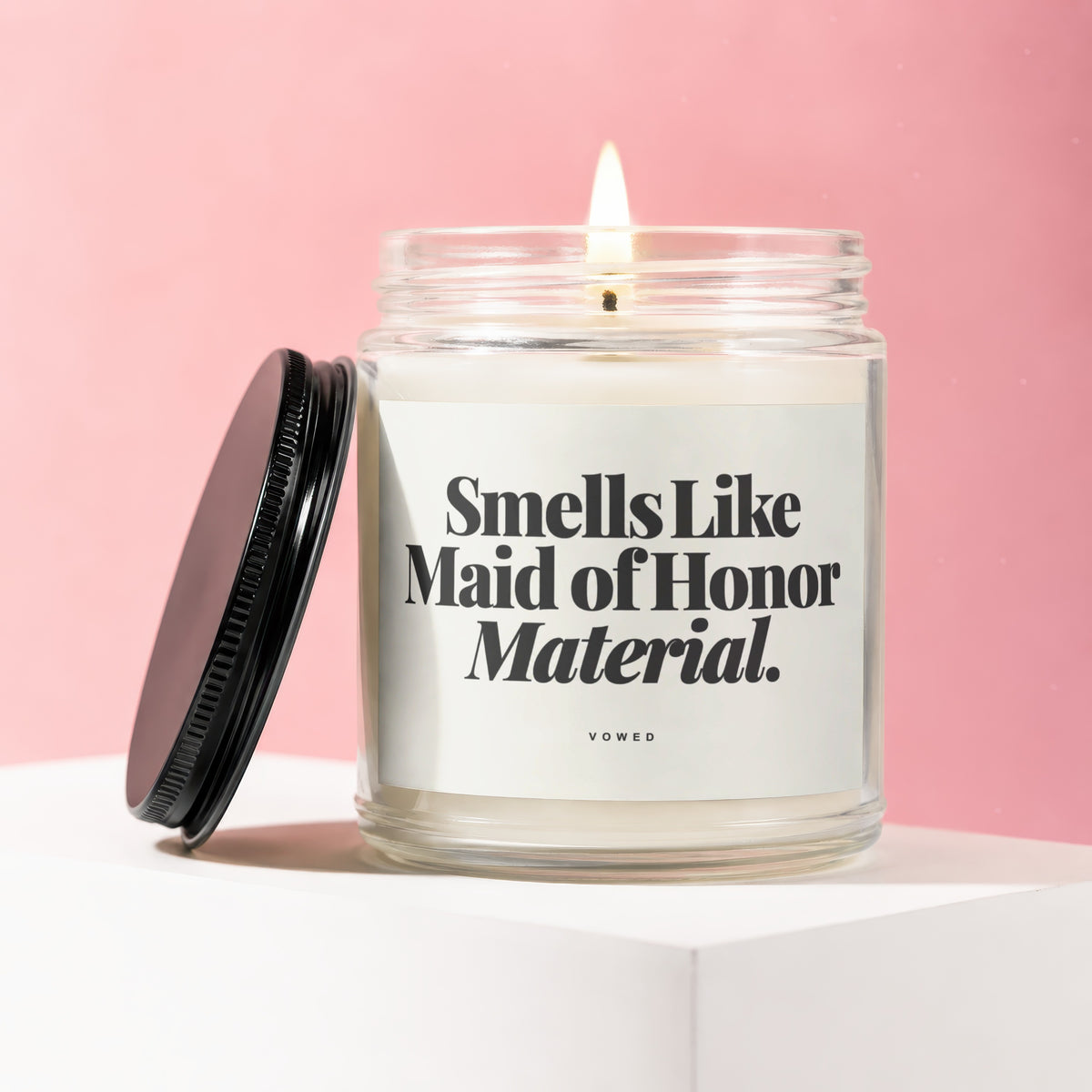 Candle with a lit wick, a black lid, and a label reading "Smells Like Maid of Honor Material. VOWED."