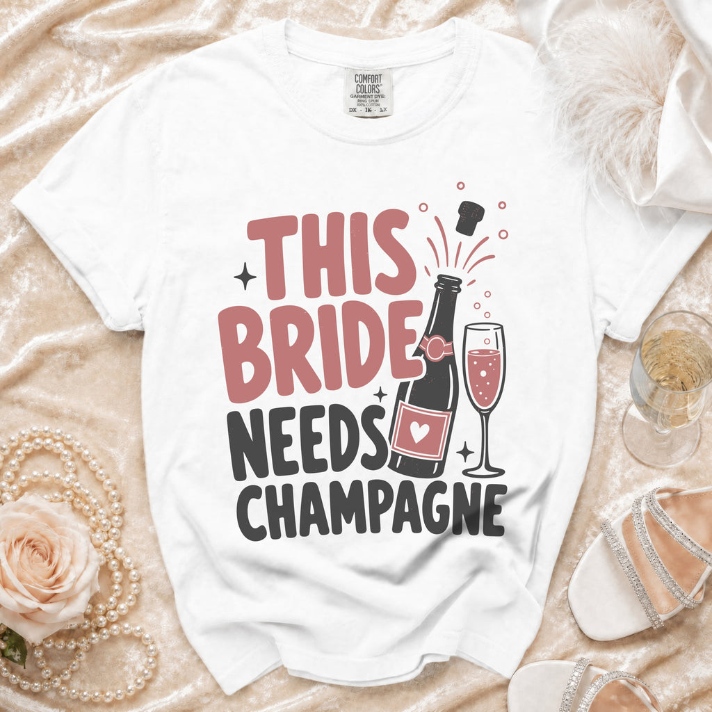 White t-shirt with "THIS BRIDE NEEDS CHAMPAGNE" text, bottle and champagne glass graphic.