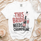 White t-shirt with "THIS BRIDE NEEDS CHAMPAGNE" text, bottle and champagne glass graphic.