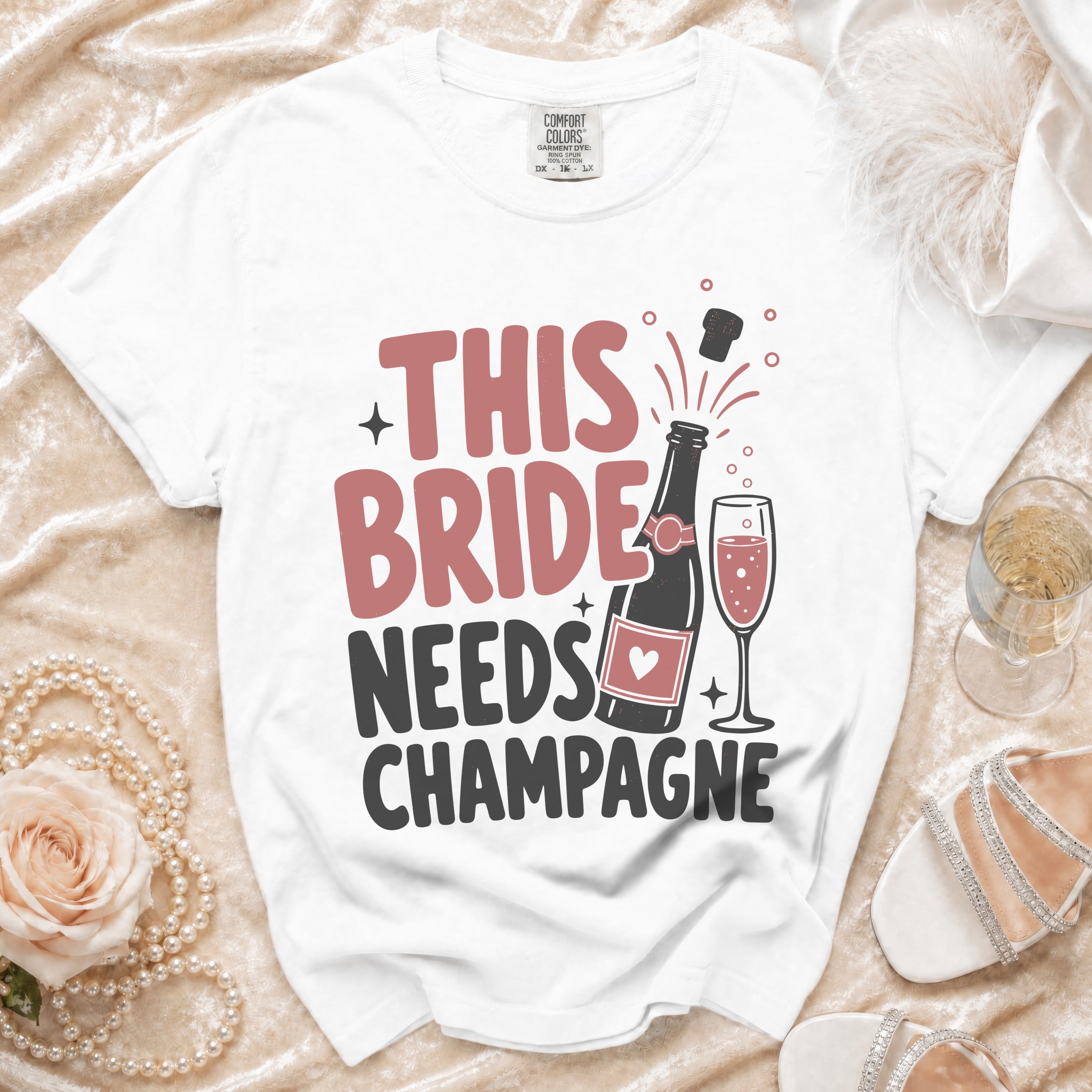 White t-shirt with "THIS BRIDE NEEDS CHAMPAGNE" text, bottle and champagne glass graphic.