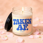 Glass candle with lit wick and a label reading "TAKEN AF. VOWED" surrounded by pink petals.