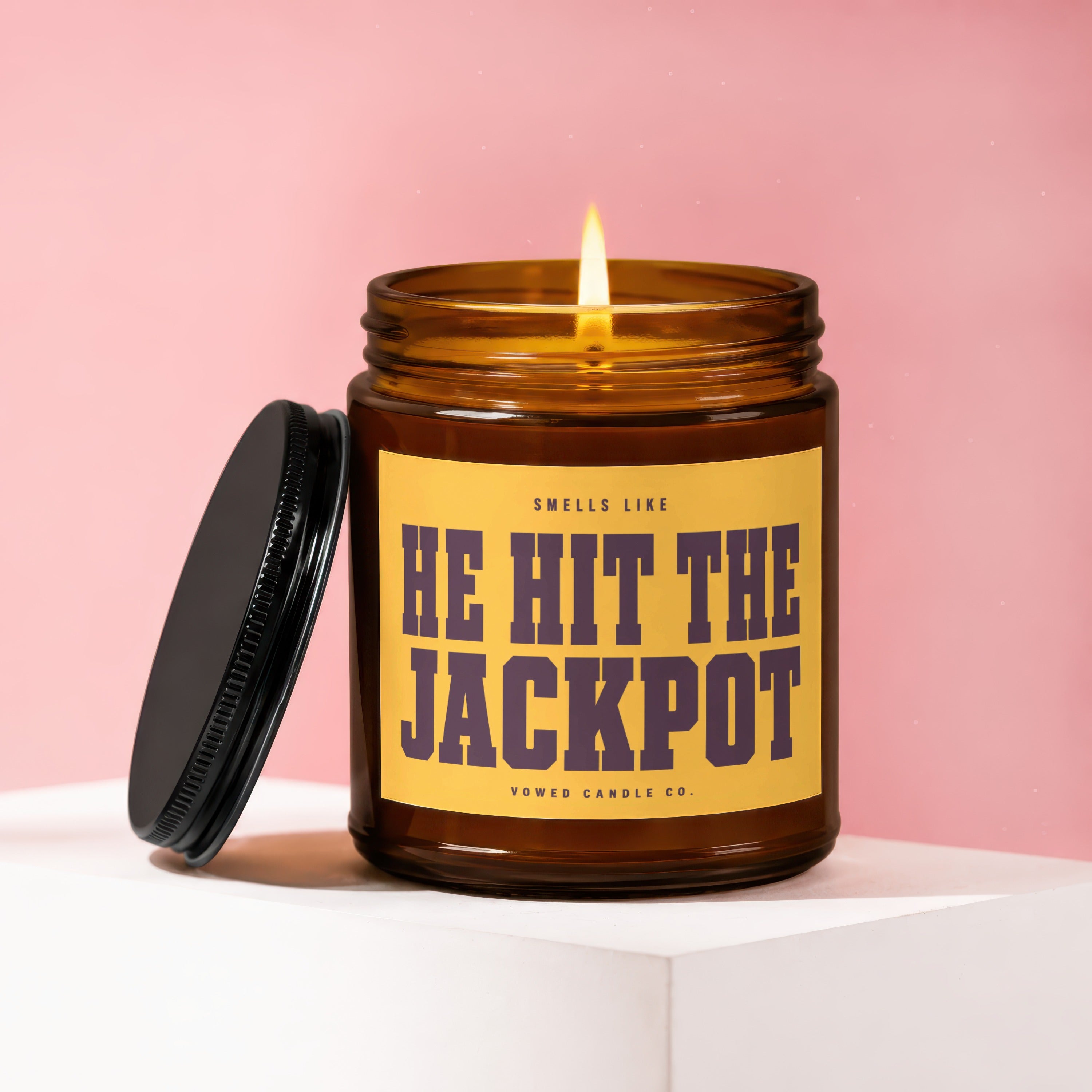 Amber jar candle with lit flame and black lid, labeled "HE HIT THE JACKPOT".
