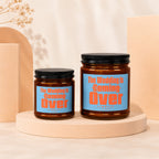 Two amber glass candles with blue labels reading "The Wedding is Coming Over" in orange.