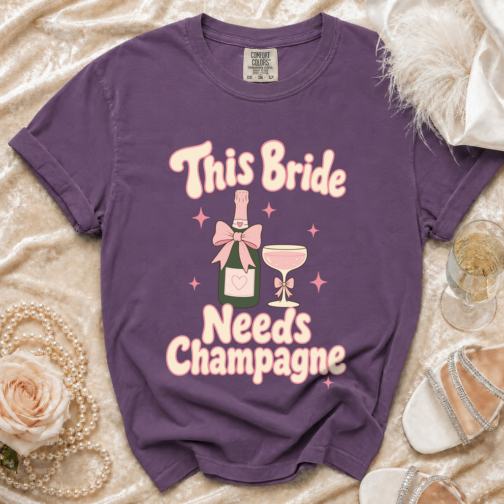 Purple t-shirt with "This Bride Needs Champagne" text, champagne bottle, and glass graphic.