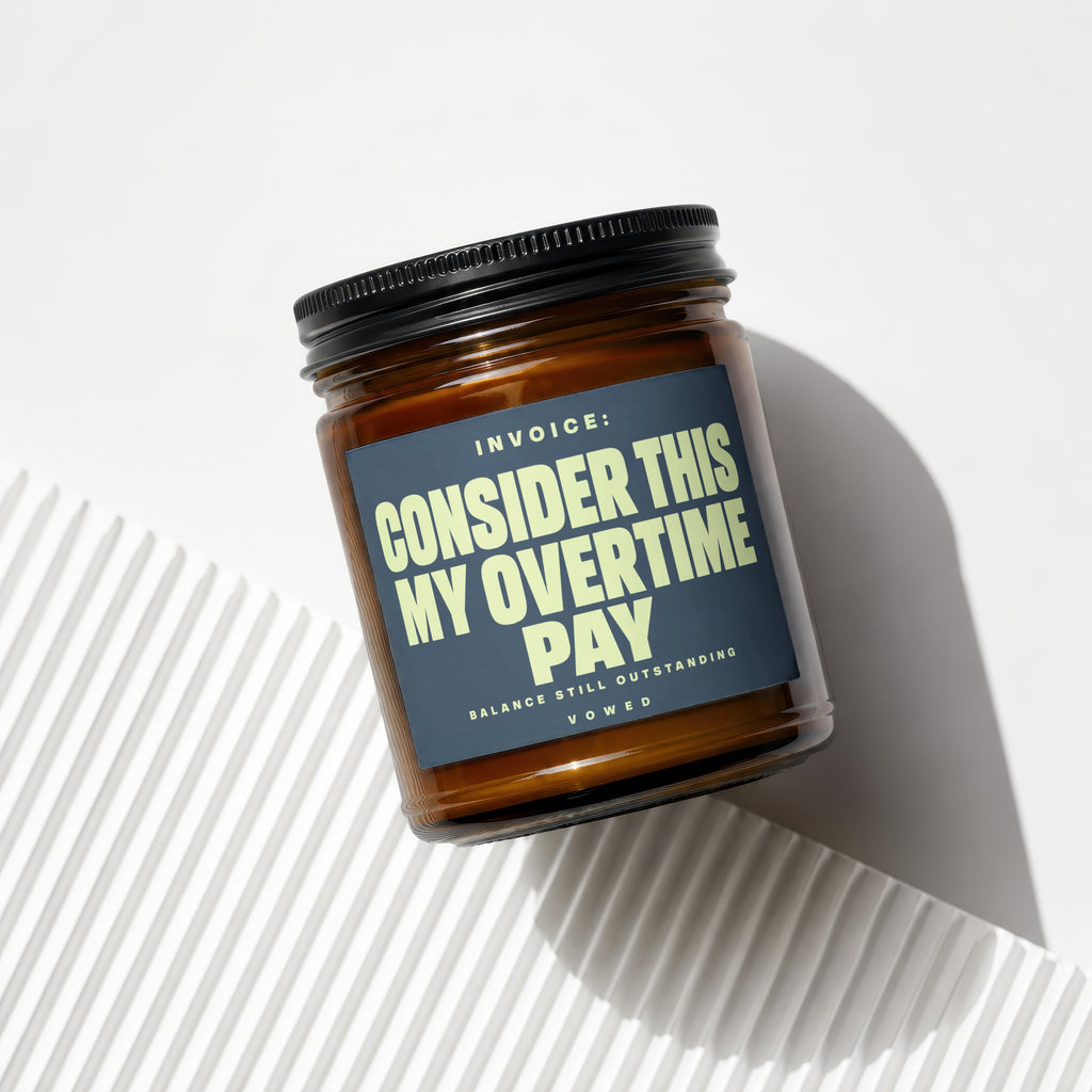 Amber glass jar candle with a dark label reading "INVOICE: CONSIDER THIS MY OVERTIME PAY".
